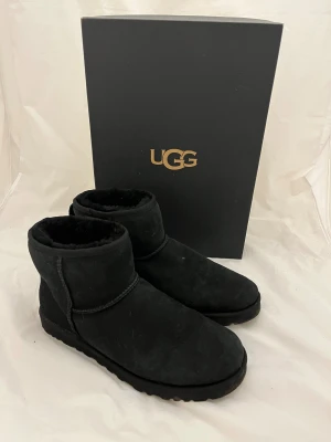 Svarta UGG boots i mocka - Klassiska svarta UGG boots i mjuk mocka med rund tå och platt sula. Insidan är fodrad med fluffig päls för extra värme och komfort. Perfekta för kyliga dagar och har en enkel, stilren design med UGG-logga bak på hälen.
