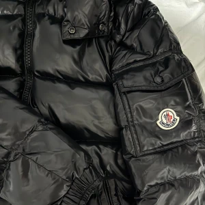 Moncler Maya  - Säljer en svart pufferjacka från Moncler med glansig finish och klassisk logga på ärmen. Jackan har dragkedja framtill, fickor med knappstängning och elastiska muddar. Insidan har coolt serietryck och autenticitetsmärkning med QR-kod. Vid intresse skriv privat för fler bilder!