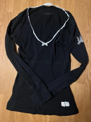 Svart ribbad långärmad tröja Odd Molly - Svart ribbad långärmad tröja från Odd Molly med vit spetskant och rosett vid v-ringningen. Snyggt tryck med fjäril på ena ärmen och liten etikett nedtill. Perfekt för dig som gillar detaljer och vill ha en unik look.