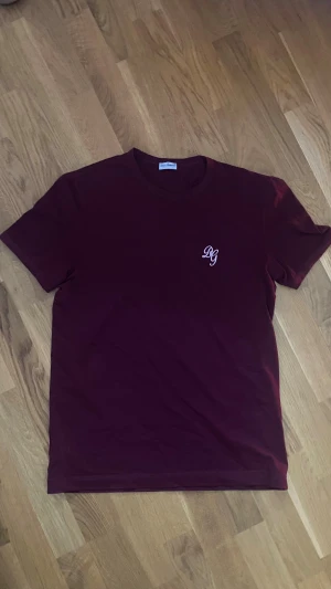 Vinröd t-shirt från Dolce & Gabbana - Snygg vinröd t-shirt från Dolce & Gabbana med broderad DG-logga på bröstet. Klassisk rund hals och korta ärmar. Tillverkad i mjuk bomull som känns skön mot huden. Perfekt för dig som gillar stilrena och exklusiva plagg.