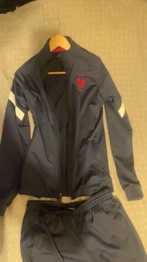 Nike FFF träningsset marinblå - Snyggt träningsset från Nike med FFF-logga, består av en marinblå zip-jacka och matchande byxor. Setet har vita detaljer på ärmar och ben samt röda broderade loggor. Materialet är lätt och syntetiskt, perfekt för sport och aktivitet.