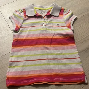 Randig pikétröja Tommy Hilfiger - Säljer en färgglad pikétröja från Tommy Hilfiger med klassisk krage och knappar framtill. Tröjan har korta ärmar och är randig i rosa, orange, röd, grön och vit. Liten broderad logga på bröstet. Perfekt för dig som gillar färg och stil!