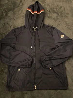 Jacka - Svart vindjacka från Moncler med huva och dragsko, hel dragkedja framtill och två fickor med tryckknappar. Jackan har Moncler-logga på ärmen och en vit, röd och marinblå rand på huvan. Tillverkad i polyester, perfekt för blåsiga dagar.