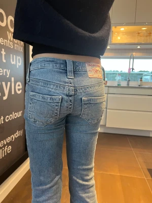 True Religion ljusblå bootcut jeans - Säljer ett par ljusblå jeans från True Religion med klassiska broderade bakfickor och bootcut passform. Jeansen har låg midja, snygga detaljer och är tillverkade i mjukt denim. Perfekt för dig som gillar ikoniska jeans med coola fickor. Modellen heter Becca Mid Rise Bootcut. Storlek 23. 