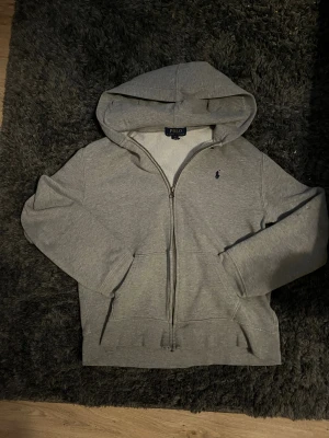 Grå hoodie Polo Ralph Lauren - Snygg grå hoodie från Polo Ralph Lauren med dragkedja och klassisk huva. Tröjan har långa ärmar, två fickor fram och den ikoniska Polo-loggan broderad på bröstet. 