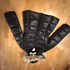 Svart pufferjacka med fuskpäls från Kappahl - Svart pufferjacka från Kappahl med avtagbar huva och fuskpäls. Jackan har bälte i midjan, dragkedja framtill och quiltad design. Perfekt för kalla dagar med sin varma och fluffiga känsla.