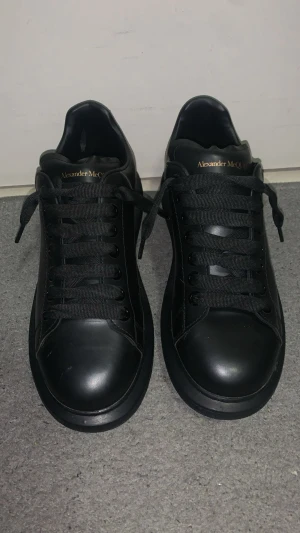 Svarta sneakers Alexander McQueen - Säljer ett par helsvarta sneakers från Alexander McQueen med chunky platt sula och rund tå. Skorna är i skinn med svarta skosnören och guldtext på häl och plös. Perfekta för dig som gillar stilrena och lyxiga sneakers med statement-känsla.