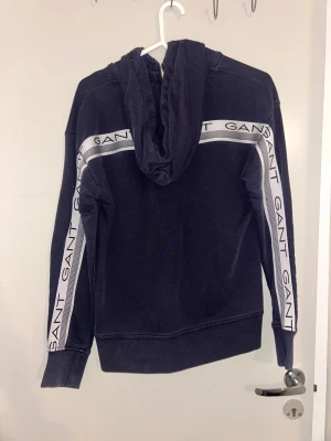 Svart hoodie med dragkedja från GANT - Svart hoodie från GANT med vit logga på bröstet och vita ränder med GANT-tryck längs ärmarna. Tröjan har huva med snörning, praktiska fickor fram och dragkedja. Perfekt för chill dagar och streetstyle.