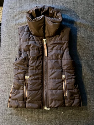Brun quiltad dunväst från JH Collection - Snygg brun dunväst från JH Collection med hög krage och quiltad design. Västen har tvåvägsdragkedja framtill, två fickor med dragkedja och bruna detaljer längs kanterna. Något sliten runt fickorna (se bilder).