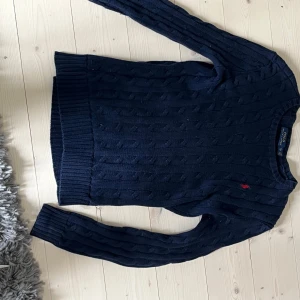 Mörkblå stickad tröja från Polo Ralph Lauren - Säljer en klassisk mörkblå stickad tröja från Polo Ralph Lauren med kabelstickat mönster och liten röd logga på bröstet. Tröjan har långa ärmar och ribbade muddar vid ärmslut och nederkant. Perfekt till många olika outfits! Har haft den ett tag och den är lite knottrig men bra skick.