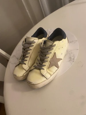 Golden Goose Superstar sneakers vit/silver - Golden Goose Superstar sneakers i vitt skinn med grå mockadetaljer och silverfärgad stjärna på sidan. Skorna har grå snörning, mörkblå hälkappa och klassisk platt sula. Snygg och ikonisk modell med cool vintagekänsla och unika detaljer.