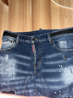 Dsquared2 blå jeans med slitningar - Snygga blå jeans från Dsquared2 med slitna detaljer, färgstänk och broderad logga i midjan. Jeansen har raka ben, knappgylf och klassisk femficksdesign. Perfekt för dig som gillar en edgy och trendig look.