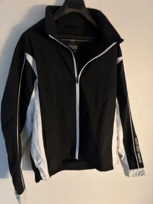 Svart och vit vindjacka (golfjacka) Galvin Green - Sportig svart och vit vindjacka från Galvin Green med coola vita paneler längs ärmarna och sidorna. Jackan har hel dragkedja framtill, hög krage och logga på kragen och ärmen. Perfekt för dig som gillar stilren och funktionell design. Storlek: L passar M. Nypris: 2999kr