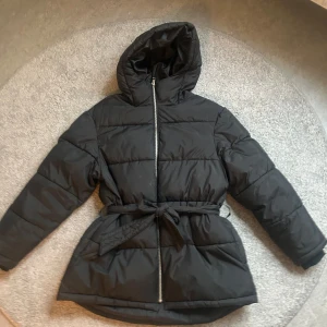 Svart dunjacka med huva - Säljer en svart dunjacka med huva och dragkedja. Jackan har ett bälte i midjan för en snygg passform och långa ärmar. Perfekt för kyliga dagar.