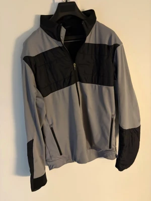 Grå och svart vindjacka (golfjacka) Galvin Green - Snygg vindjacka från Galvin Green i grått och svart med halv dragkedja framtill. Jackan har hög krage, två sidofickor med dragkedja och coola svarta paneler över bröst och armar. Perfekt för dig som gillar sportig stil och vill ha något funktionellt. Utmärkt golfjacka. Storlek: L passar M. Nypris: 2000kr