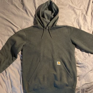 Grå Carhartt hoodie - Säljer en grå hoodie från Carhartt  med huva och snörning. Tröjan har en stor magficka och klassisk Carhartt-logga framtill. Perfekt för chill dagar och streetwear-stil. Materialet är mjuk bomull och polyesterblandning, vilket gör den väldigt mjuk och len
