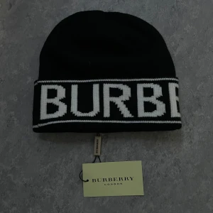 Burberry mössa - Svart mössa från Burberry med bred vit logotyp runt uppviket. Stilren och klassisk design som ger en cool vibe. Materialet är stickat och känns mjukt mot huden. Perfekt för att lyfta din streetstyle under kyliga dagar.