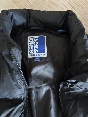 Svart pufferjacka från Jack & Jones - Säljer en svart pufferjacka från Jack & Jones i storlek 164. Jackan har quiltad design, hög krage och dragkedja framtill. 