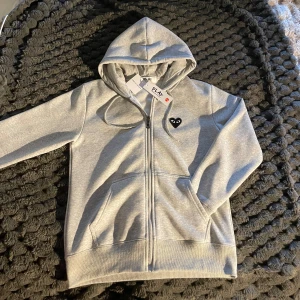Grå hoodie Comme des Garçons Play - Grå hoodie från Comme des Garçons Play med dragkedja, huva och snörning. Ikoniskt svart hjärta med ögon på bröstet och stort tryck på ryggen. Mjuk bomullskvalitet och kängurufickor framtill. Perfekt för en chill och stilren look. PRIS KAN DISKUTERAS