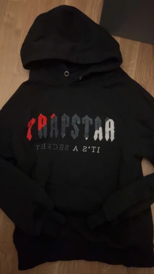 Trapstar svart hoodie med tryck - Svart hoodie från Trapstar med stor huva och tryck i rött, grått och vitt på bröstet. Tröjan har långa ärmar och ribbade muddar. Materialet är mjuk bomull och passformen är normal. Perfekt för dig som gillar streetwear och vill sticka ut.