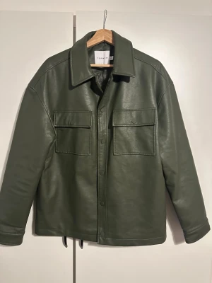 Grön skinnovershirt från Topman - Snygg mörkgrön overshirt från Topman i skinnimitation med quiltat foder. Jackan har två stora bröstfickor med lock, tryckknappar framtill och klassisk krage. Perfekt för dig som gillar stilren och modern look. 