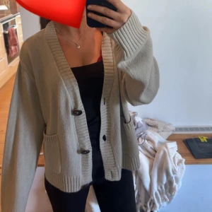 Beige stickad kofta med knappar - Säljer en beige stickad kofta med två stora knappar framtill och bred ribbad kant vid hals och ärmslut. Koftan har två praktiska fickor och en loose passform med långa ärmar. Perfekt att slänga över en topp för en chill look.