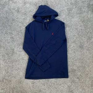 Marinblå hoodie från Polo Ralph Lauren - Snygg marinblå hoodie från Polo Ralph Lauren med klassisk röd logga broderad på bröstet. Tröjan har huva med snörning och är tillverkad i mjuk bomull. Perfekt för en chill och stilren look.