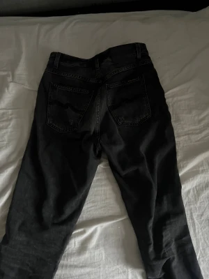 Svarta jeans från Nudie Jeans, 27/30 - Svarta jeans från Nudie Jeans med klassisk femficksdesign och raka ben. Jeansen har subtila sömdetaljer på bakfickorna och orange broderad logga på insidan. Tillverkade i mjukt denimtyg som sitter snyggt och passar till allt.