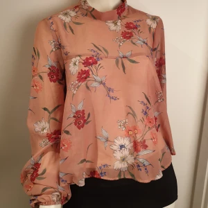 Blommig blus från Gina Tricot - En söt blommig blus från Gina Tricot i en mjuk persikofärg. Blusen har långa ärmar och ett knytband i nacken. Perfekt för en romantisk look med sitt vackra blommönster i rött, vitt och blått.