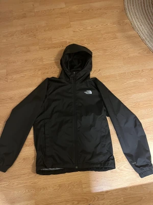 The North Face jacka  - Svart vindjacka från The North Face med huva och dragkedja framtill. Jackan har två sidofickor, elastiska muddar och meshfoder på insidan. Perfekt för blåsiga dagar och enkel att matcha med allt. Klassisk logga på bröstet och ryggen. Man kan enkelt spänna åt i midjan.