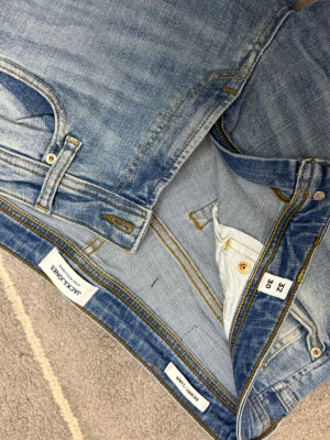 Blåa Jack&Jones jeans  - Säljer ett par ljusblå skinny jeans från Jack & Jones, modell Skinny Liam. Jeansen har klassisk femficksdesign, gul kontrastsöm och knappgylf. Materialet är mjukt denim med snygga slitningar och tvättade detaljer för en cool look.