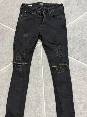 Svarta slim fit jeans Jack & Jones - Svarta slim fit jeans från Jack & Jones, modell Glenn. Jeansen har slitna detaljer och dragkedjor vid knäna för en edgy look. Klassisk femficksdesign och knappgylf. Perfekta för dig som gillar streetstyle och vill sticka ut.