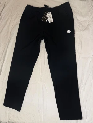 Svarta byxor från Descente - Svarta byxor från Descente med elastisk midja och snörning. Byxorna har raka ben och en vit logga på vänster lår. Sköna fickor på sidorna och mjukt material som passar chill och sport. Perfekt för dig som gillar enkel och stilren design.