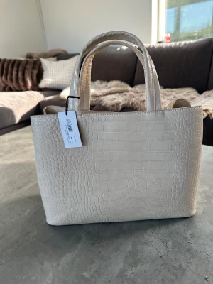 Beige handväska i croco från NA-KD - Stilren beige handväska från NA-KD med croco-mönstrad yta och två rejäla handtag. Väskan har flera fack, inklusive ett med dragkedja, och stängs med magnetknapp. Perfekt för dig som vill ha en trendig och rymlig väska.