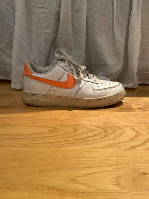 Nike Air Force 1 vit och orange sneaker - Nike Air Force 1 sneakers i vitt skinn med orange swoosh och häl. Låg modell med snörning och platt sula. Insidan har orange mesh-detaljer och grått foder. Klassisk rund tå och tydlig Air-logga på hälen. Perfekt för dig som gillar retrovibe och färg.