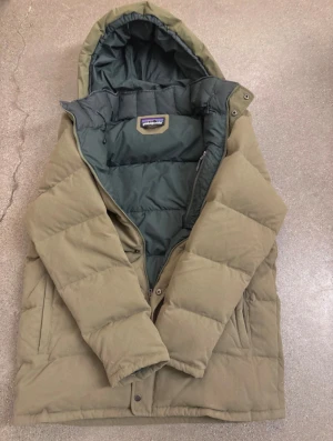  Patagonia pufferjacka - Säljer en olivgrön dunjacka från Patagonia med huva och quiltad design. Jackan har dragkedja och tryckknappar framtill, två sidofickor och en innerficka med dragkedja. Perfekt för kalla dagar och har en clean, stilren look.