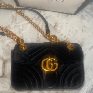 Svart sammet axelväska från Gucci - Supersnygg svart axelväska från Gucci i sammet med guldiga kedjedetaljer och GG-logo framtill. Väskan har quiltad design med hjärtdetalj på baksidan och dragkedja inuti. Perfekt för dig som vill sticka ut med lyxig känsla och cool vibe.