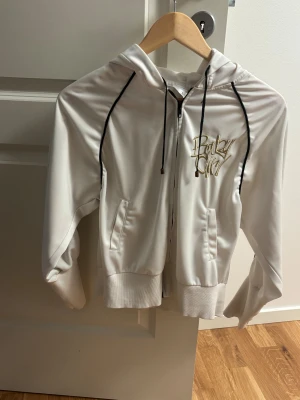 Vit hoodie Baby Girl by Terranova - Vit hoodie med broderad Baby Girl-text i guld på bröstet. Svarta snören och svarta ränder längs ärmarna ger en sportig vibe. Dragkedja framtill och två fickor. Mjuk insida och ribbade muddar. Perfekt för chill dagar.