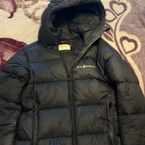 Svart dunjacka från Sail Racing - Svart dunjacka från Sail Racing med huva och dragkedja. Jackan har en puffig design, logga på bröstet och huvan samt två sidofickor. Perfekt för kalla dagar och riktigt snygg streetstil.