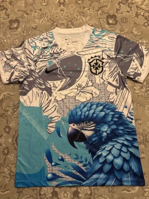 Brazil 25/26 specialutgåva - Unik fotbollströja från Nike med brazil-logga, korta ärmar och Dri-FIT-material. Designen har ett stort blått papegojmotiv, blommor och grafiska detaljer i blått, vitt och grått. Perfekt för dig som gillar färgstarka och kreativa sportplagg.