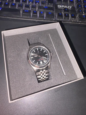 Svart och silverfärgad klocka från GANT - Stilren klocka från GANT med svart urtavla, datumvisning och silverfärgad länk i rostfritt stål. Klassisk rund form med räfflad bezel och tydliga index. Perfekt accessoar för dig som gillar en clean look och vill ha koll på tiden.