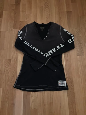 Svart långärmad tröja True Religion - Svart långärmad tröja från True Religion med vit text längs ärmarna och coola detaljer som v-ringning med knappar. Materialet är 100% bomull med ribbad struktur och patch nertill. Snygga kontrastsömmar och street-inspirerad look. Från 2008, inga defekter! 