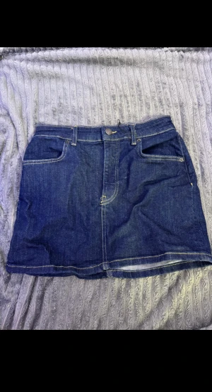 Mörkblå jeanskjol med fickor - Snygg mörkblå jeanskjol med klassiska fickor både fram och bak, samt bälteshällor och synliga kontrastsömmar. Kjolen har en kort och rak passform med knapp och dragkedja framtill. Perfekt för dig som gillar enkel denimstil.