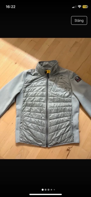 Ljusgrå quiltad jacka Parajumpers - Snygg ljusgrå jacka från Parajumpers med quiltad framsida och detaljer i mesh. Jackan har dragkedja, hög krage med gul knappdetalj och patch på ärmen. Perfekt för dig som gillar sportig stil och vill ha något som sticker ut.