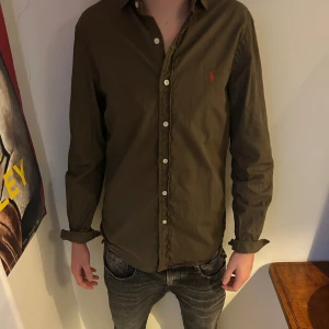 Olivgrön skjorta från Ralph Lauren - Snygg olivgrön skjorta från Ralph Lauren med klassisk krage och knappar hela vägen. Den har lång ärm och den ikoniska röda Polo-loggan broderad på bröstet. Skjortan är i mjuk bomull och har en normal passform som funkar till jeans eller chinos.