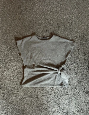 Grå topp med knyt från Zara - Säljer en grå topp från Zara med snygg knytning i sidan. Toppen har en enkel, rak passform och är ärmlös med rund halsringning. Perfekt för en chill och stilren look. Materialet känns mjukt och bekvämt mot huden.