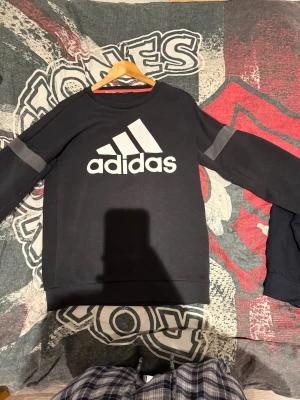 Svart Adidas sweatshirt med logga - Svart sweatshirt från Adidas med stor vit logga på bröstet och gråa detaljer på ärmarna. Tröjan har rund hals och långa ärmar, perfekt för chill eller träning. Materialet är mjuk bomull och polyesterblandning som känns skönt mot huden.