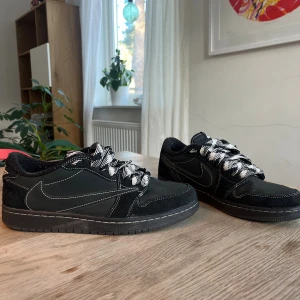 Nike Air Jordan 1 Low Travis Scott - Svarta Nike Air Jordan 1 Low Travis Scott med vita kontrastsömmar och unika svartvita snören. Skorna har detaljer i mocka och skinn, broderad Air Jordan-logga och bi på hälen. Snygg och ikonisk siluett med rund tå och platt sula. Använda fåtal gånger men skulle säga att de fortfarande är i väldigt bra skick. 