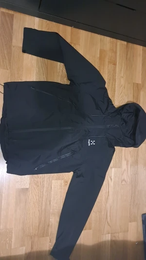 Svart vindjacka från Haglöfs med huva - Svart vindjacka från Haglöfs med snygg minimalistisk design, justerbar huva och flera dragkedjeförsedda fickor. Jackan är tillverkad i slitstark polyester och har vattentäta dragkedjor. Perfekt för dig som gillar att vara ute oavsett väder. Storlek S men passar M helt ny med etikett på 