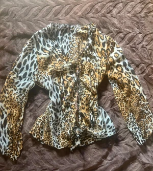 Leopardmönstrad blus med snörning - Säljer en vild leopardmönstrad blus med coola snörningsdetaljer framtill och längs midjan. Blusen har långärmade, lätt puffade ärmar och är gjord i ett tunt, flowy material som känns riktigt skönt. Färgerna går i beige, brun, svart och vit med kontrasterande mönster.
